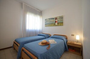 Ein Zimmer mit zwei Einzelbetten, einem Nachttisch mit Lampe, einem Gemälde von einem Leuchtturm und einem Fenster mit weißen Vorhängen.