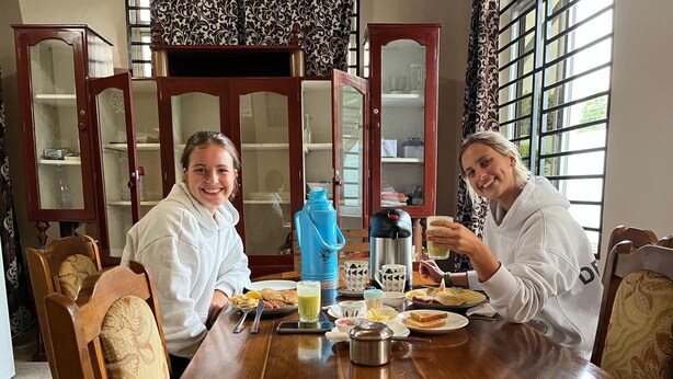 Dos personas sentadas en una mesa de madera con una botella de agua azul, una cafetera y diversos alimentos para el desayuno.