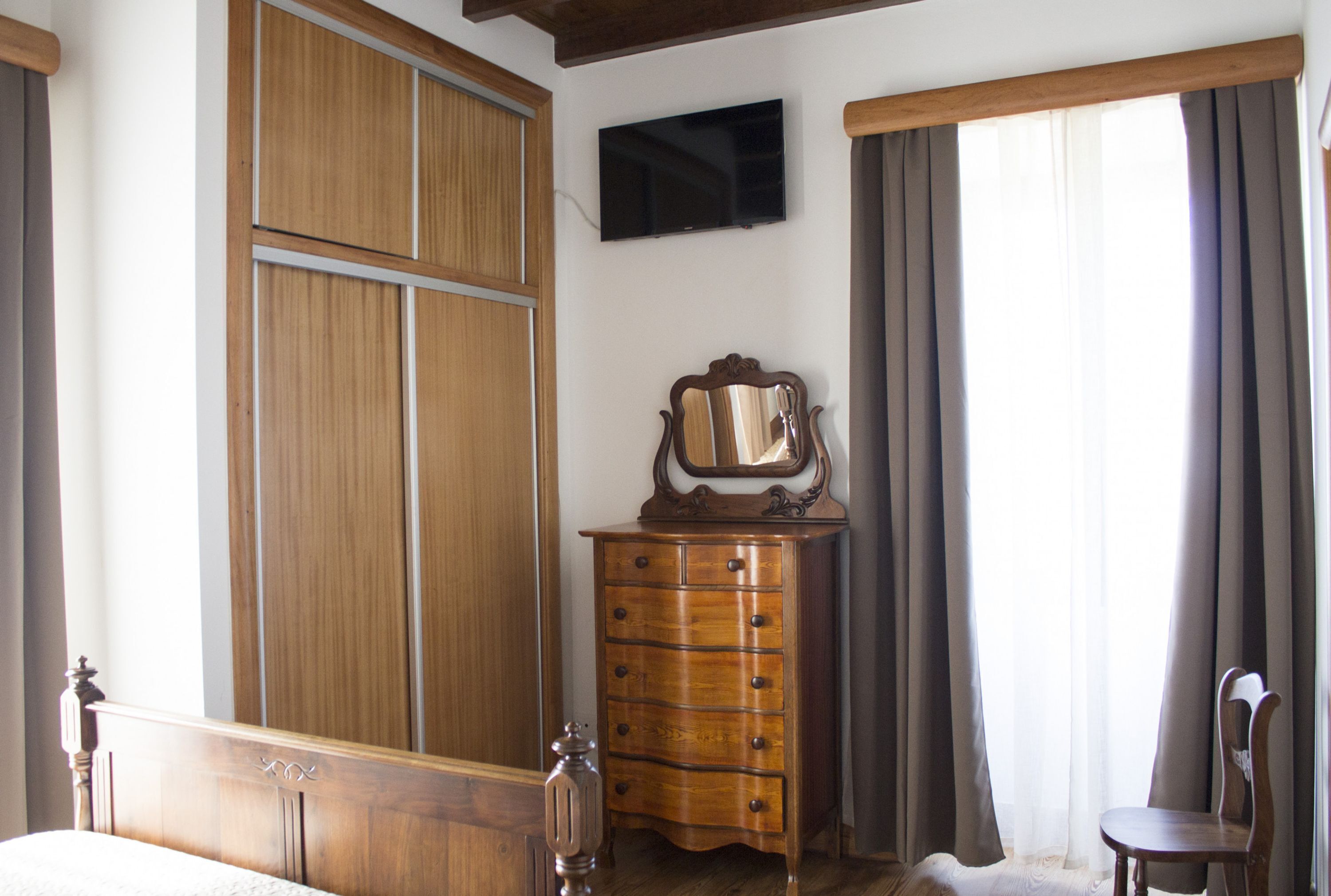 Ein Schlafzimmer mit einem hölzernen Bett, einem Schminktisch, einem Spiegel, einem an der Wand befestigten Fernseher und einem fenster mit Vorhängen.