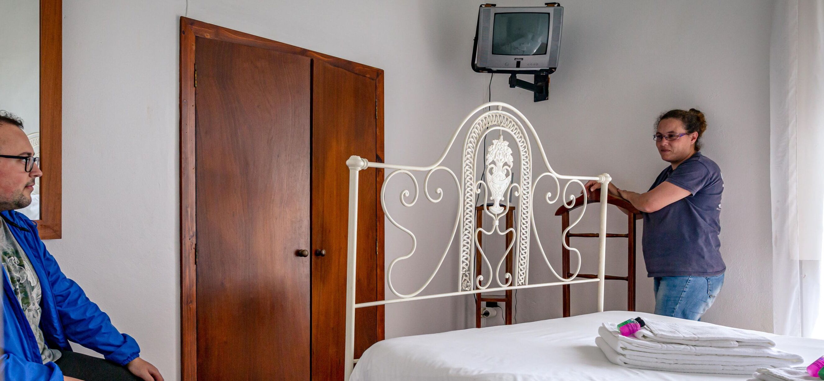 Quarto com guarda-roupa de madeira, estrutura de cama de metal branco, televisão montada na parede e cama com roupa de cama branca e bem arrumada.