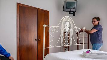 Quarto com guarda-roupa de madeira, estrutura de cama de metal branco, televisão montada na parede e cama com roupa de cama branca e bem arrumada.