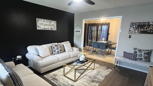 Ein Wohnzimmer mit einem weißen Sofa, einem Couchtisch und einem Deckenventilator. Durch eine offene Tür ist eine Essecke erkennbar.