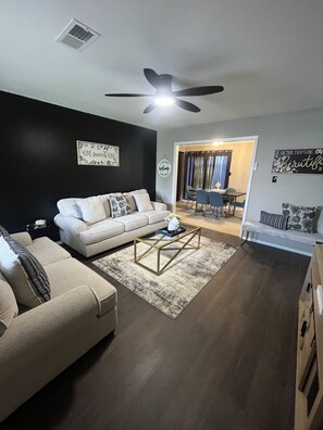 A living room with a white sofa, a coffee table, and a ceiling fan. There is a dining area visible through an open door. - The 'Serene' Retreat has everything you need. You will feel like this is HOME! (Greenville)