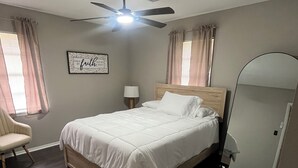 Ein Schlafzimmer mit einem Bett, einem Sessel, einem Deckenventilator, einer Nachttischlampe und einem Spiegel.
