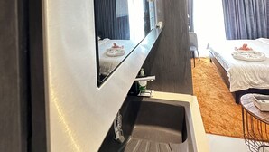 A modern hotel room with a large flat-screen TV mounted on the wall, a built-in sink, a bed with a white comforter, and a small round table with a chair. - Easy Stay at the Lennox  (Accra)