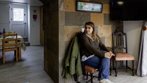 Une personne est assise sur une chaise en bois dans une pièce dont un mur est en pierre. Un téléviseur est fixé au mur, et on aperçoit un coin repas à l’arrière-plan.