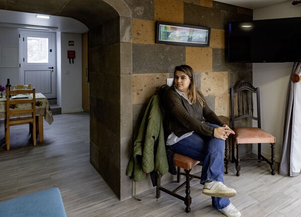 Uma pessoa sentada em uma cadeira de madeira em um quarto com parede de pedra, uma televisão fixada na parede, e uma área de jantar visível ao fundo.