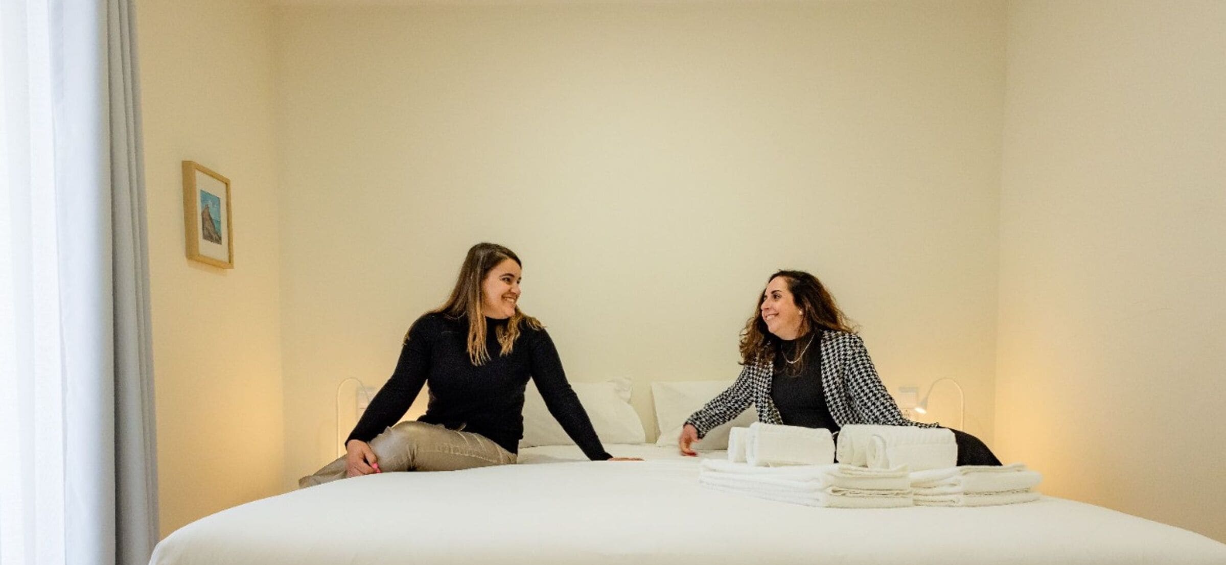 Ein Hotelzimmer mit einem Bett, zwei Personen darauf sitzend und einem Stapel Handtücher auf dem Bett.