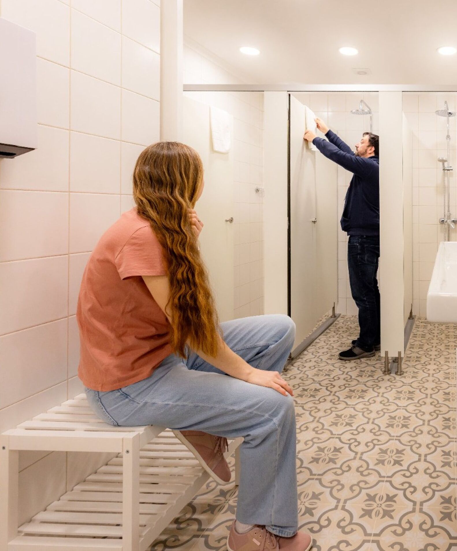 Baño con un banco, una persona sentada y otra de pie frente a un espejo.