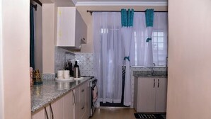 A compact kitchen with white cabinets, a granite countertop, and a window with white curtains. - Lux Suites comfy Homestays Nakuru (Nakuru)