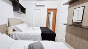 A hotel room with two beds, a bedside table with a lamp, a mirror, a shelf with a plant, and an air conditioning unit. - COPIT0105 - Edifício Cidade de Caravelas (Salvador)