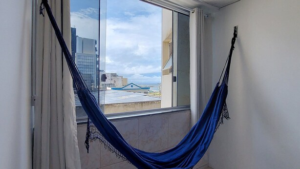 A room with a window, a blue hammock, and a view of buildings and a blue structure outside. - COPIT0105 - Edifício Cidade de Caravelas (Salvador)