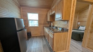 A compact kitchen with wooden cabinets, a black refrigerator, and a small window. - Wichita's Spring Lake RV Resort (Halstead)
