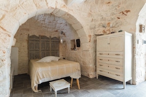 A bedroom with a stone archway, a bed with a patterned bedspread, a wooden nightstand, a dresser, and a small table. - Trullo Letizia con Piscina by Wonderful Italy (Ostuni)