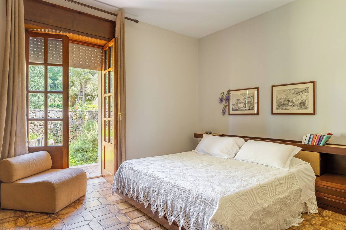 Ein Schlafzimmer mit Bett, Stuhl, einem Holzkopfstück und Blick auf Grün hinter einem Fenster.