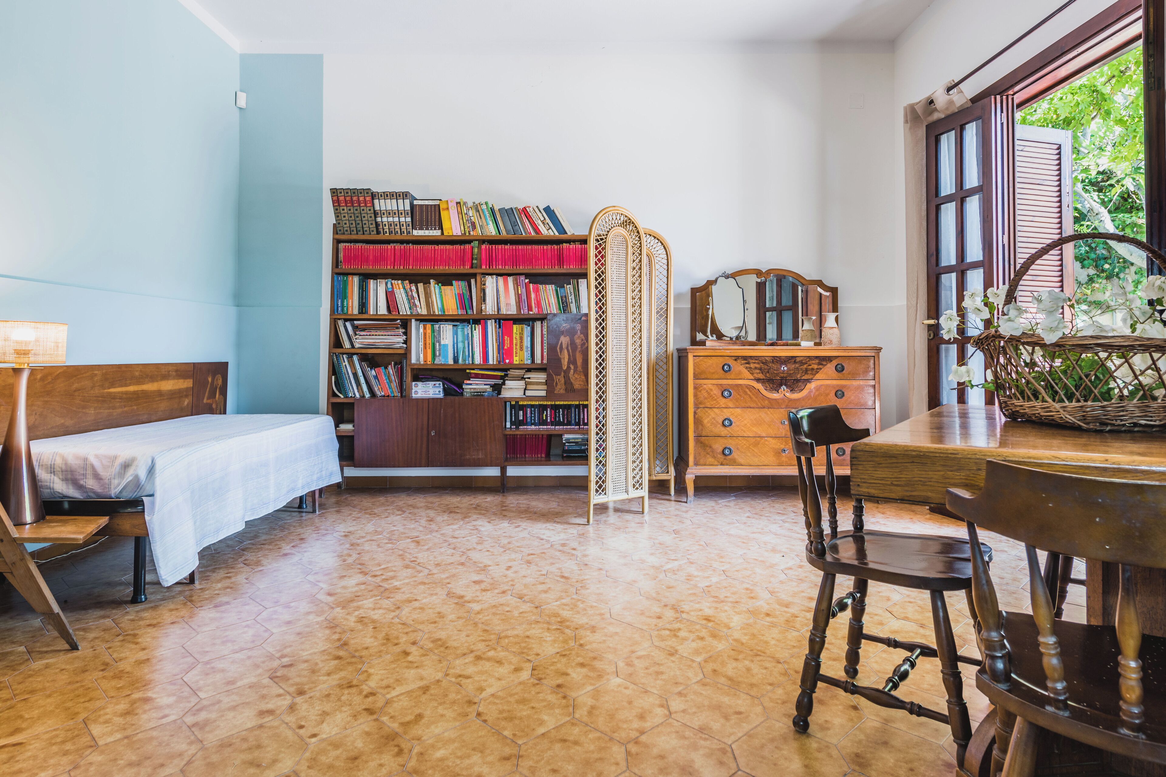 A cozy room with a bed, a wooden dresser, a bookshelf, and a window with a view of greenery.