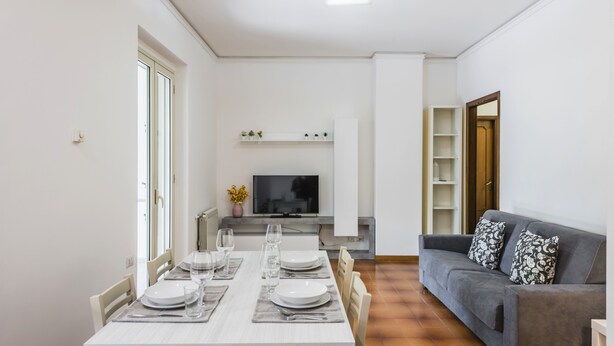 Een moderne woonkamer met een eetgedeelte, een grijze bank en een televisie op een dressoir.