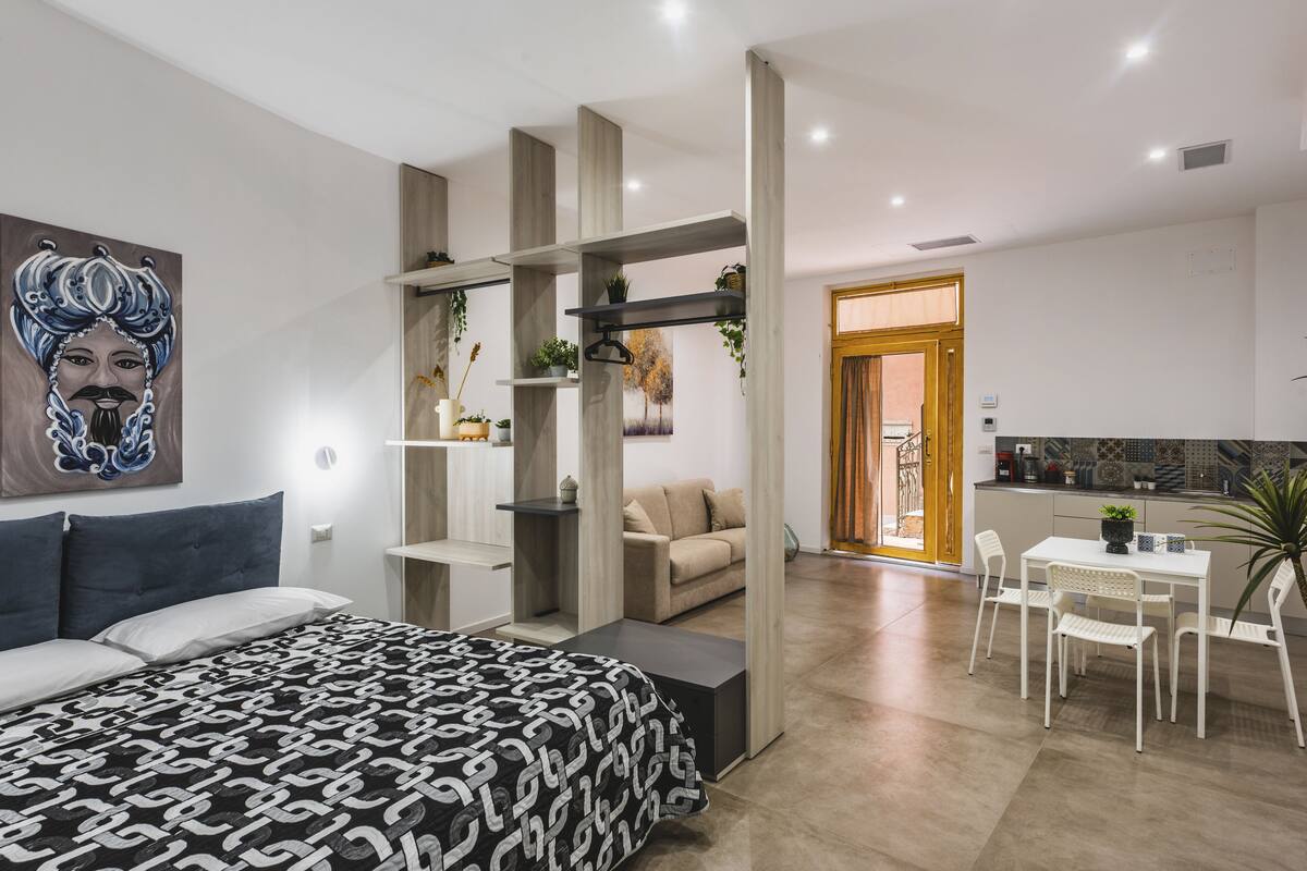 Ein modernes Schlafzimmer mit Bett, Schreibtisch und Essbereich. An der Wand hängt ein Bild, eine Pflanze steht daneben und es gibt einen Blick ins Freie.