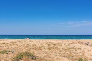 A beach with two people sitting on the sand, a clear blue sky, and the ocean in the background. - Il Cancello sul Mare by Wonderful Italy (Avola)