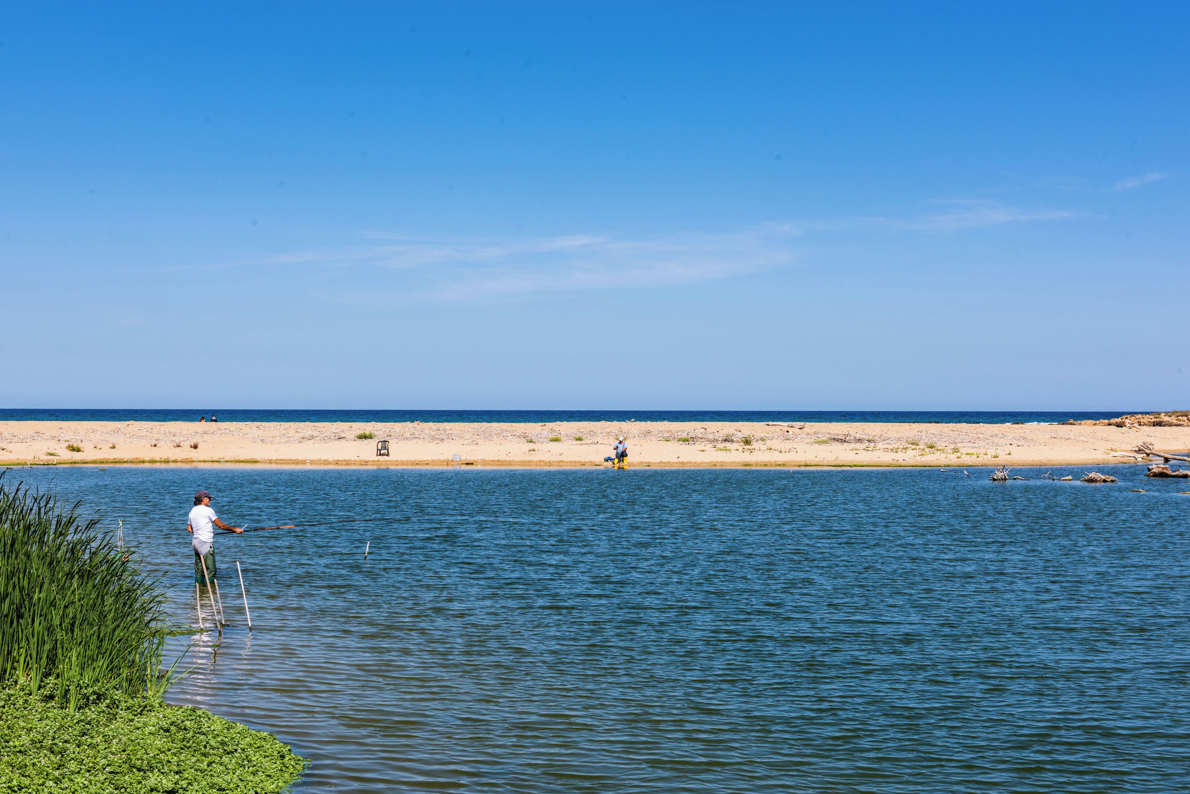 Une personne est en train de pêcher au bord d’une étendue d’eau calme, avec une plage de sable en arrière-plan.