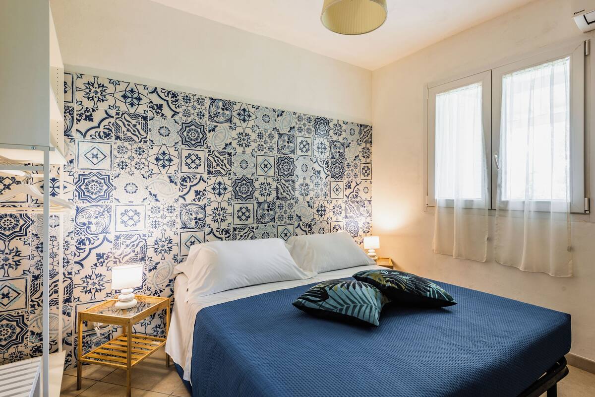 A bedroom with a patterned blue and white tile wall, a bed with white and blue bedding, a bedside table with a lamp, and a window with sheer curtains.
