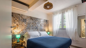 A bedroom with a patterned tile wall, a bed with a blue bedspread, a bedside table with a lamp, and a window with sheer curtains. - Il Cancello sul Mare by Wonderful Italy (Avola)