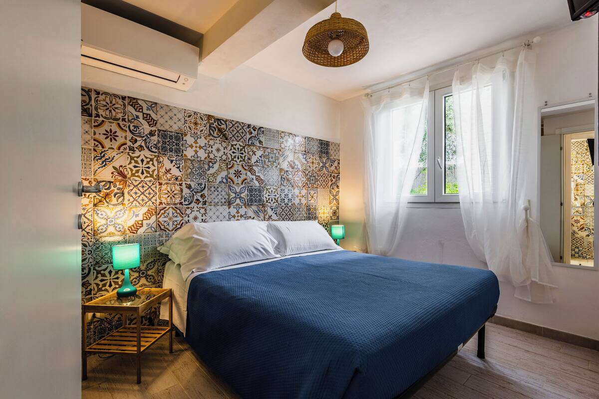 A bedroom with a patterned tile wall, a bed with a blue bedspread, a bedside table with a lamp, and a window with sheer curtains.