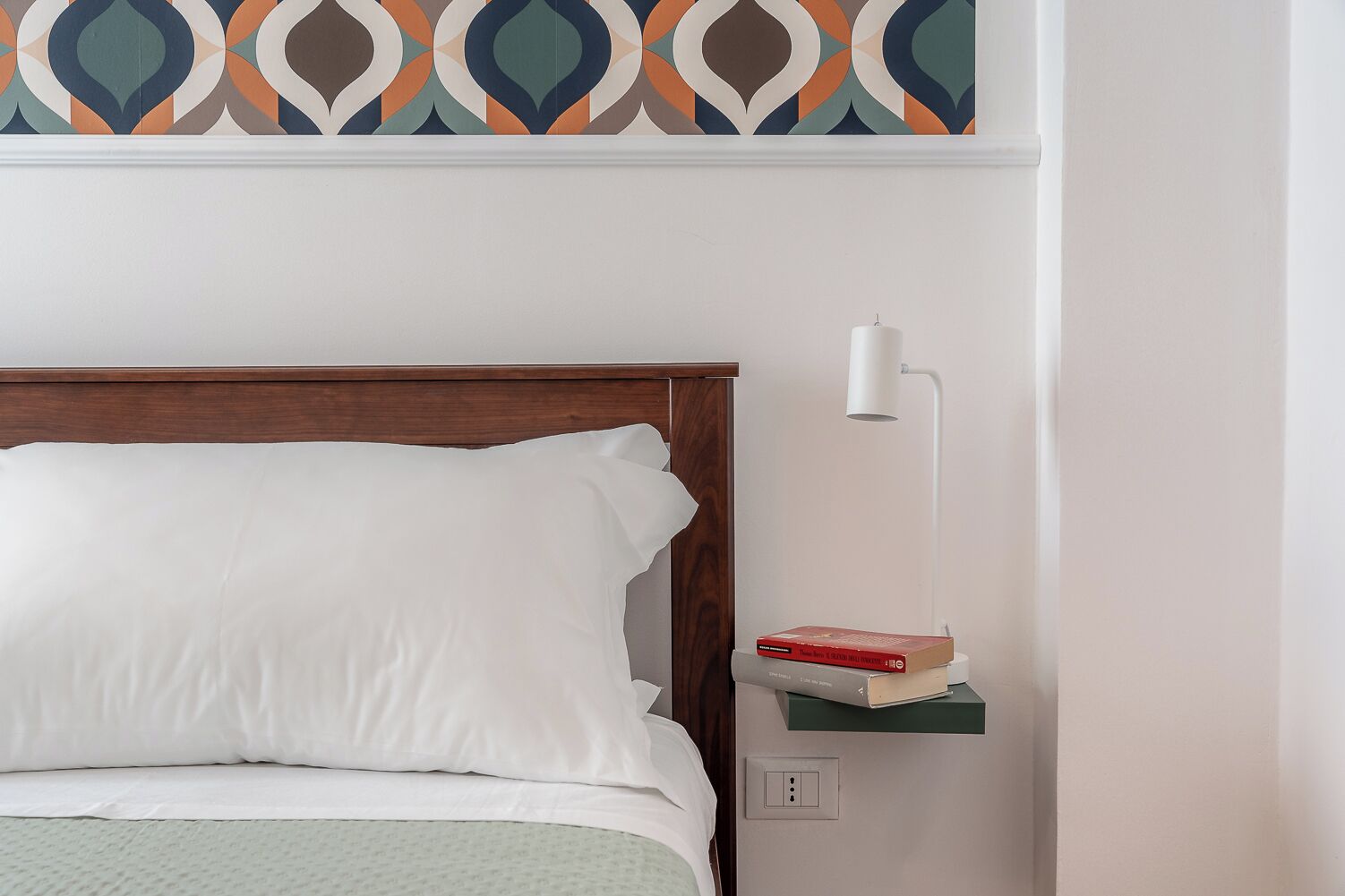 A neatly made bed with white linens, a wooden headboard, a bedside lamp, and a small shelf with books.