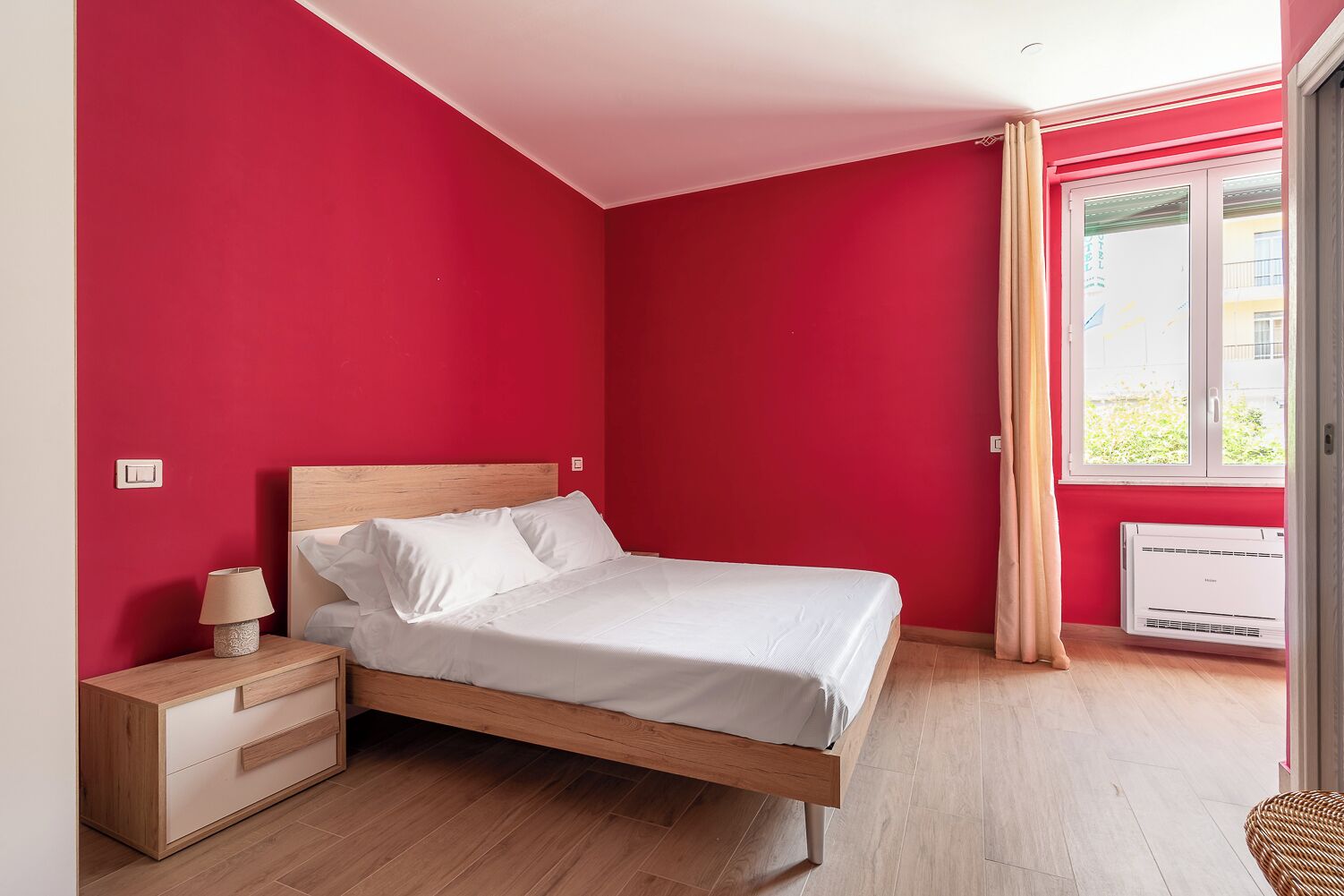 A bedroom with a bed, wooden nightstand, lamp, and an air conditioning unit.