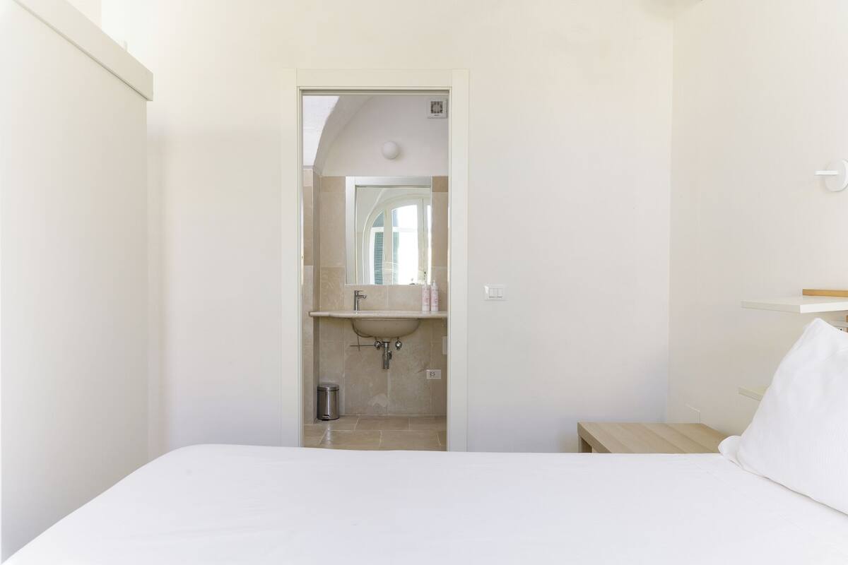 Un dormitorio con cama, mesita de noche, espejo sobre el lavamanos y una puerta que da al baño.