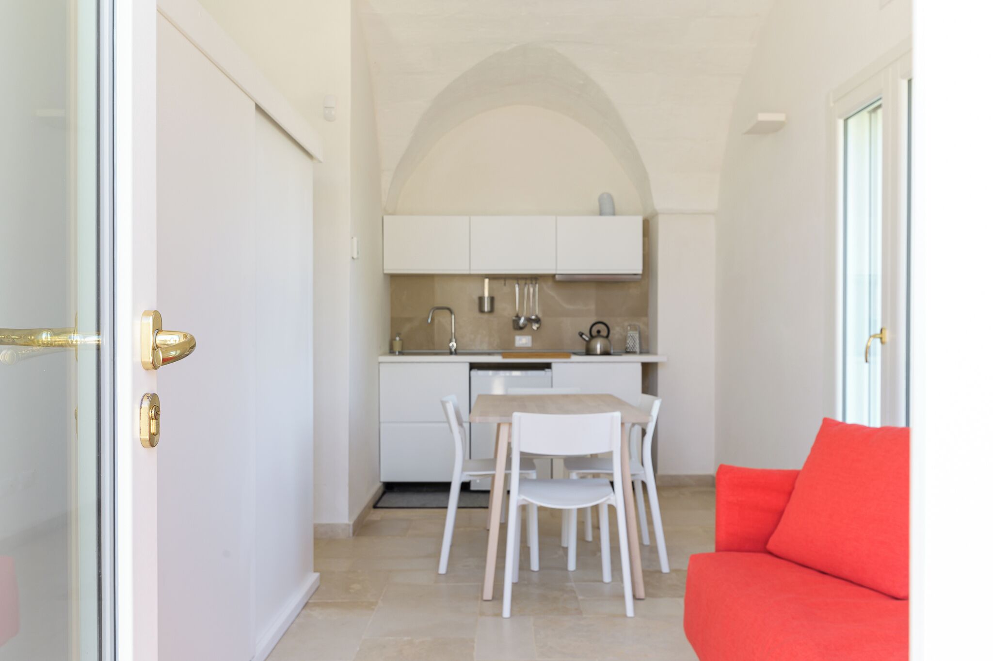 Une cuisine compacte avec des meubles blancs, une table à manger avec des chaises et un canapé rouge dans une pièce lumineuse dotée d’un sol carrelé.