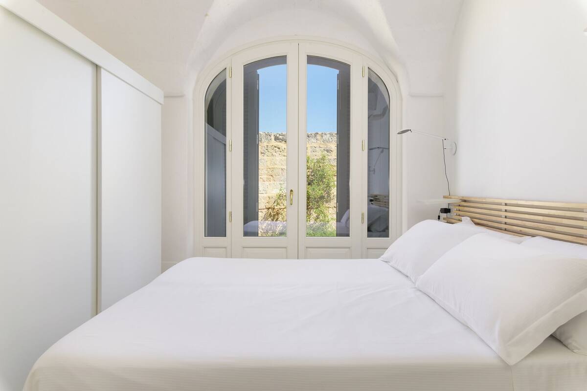 Un dormitorio con cama, paredes blancas, cabecera de madera y vista a un paisaje rocoso a través de una puerta de cristal.