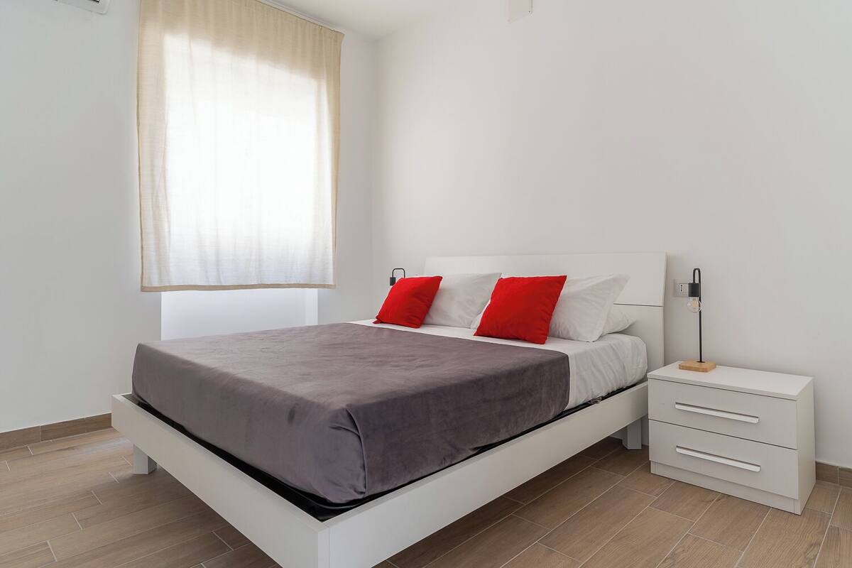 Un dormitorio moderno con cama, mesitas de noche blancas y una ventana con cortinas traslúcidas.