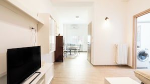 Ein modernes Apartment mit einem Flachbildfernseher auf einem weißen Standfuß, einer Essecke mit Tisch und Stühlen, einer Waschmaschine und einem Heizkörper.