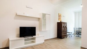 Ein modernes Wohnzimmer mit einem Flachbildfernseher auf einer weißen Unterhaltungseinheit, einem Holzschrank und einem Essbereich mit Tisch und Stühlen.