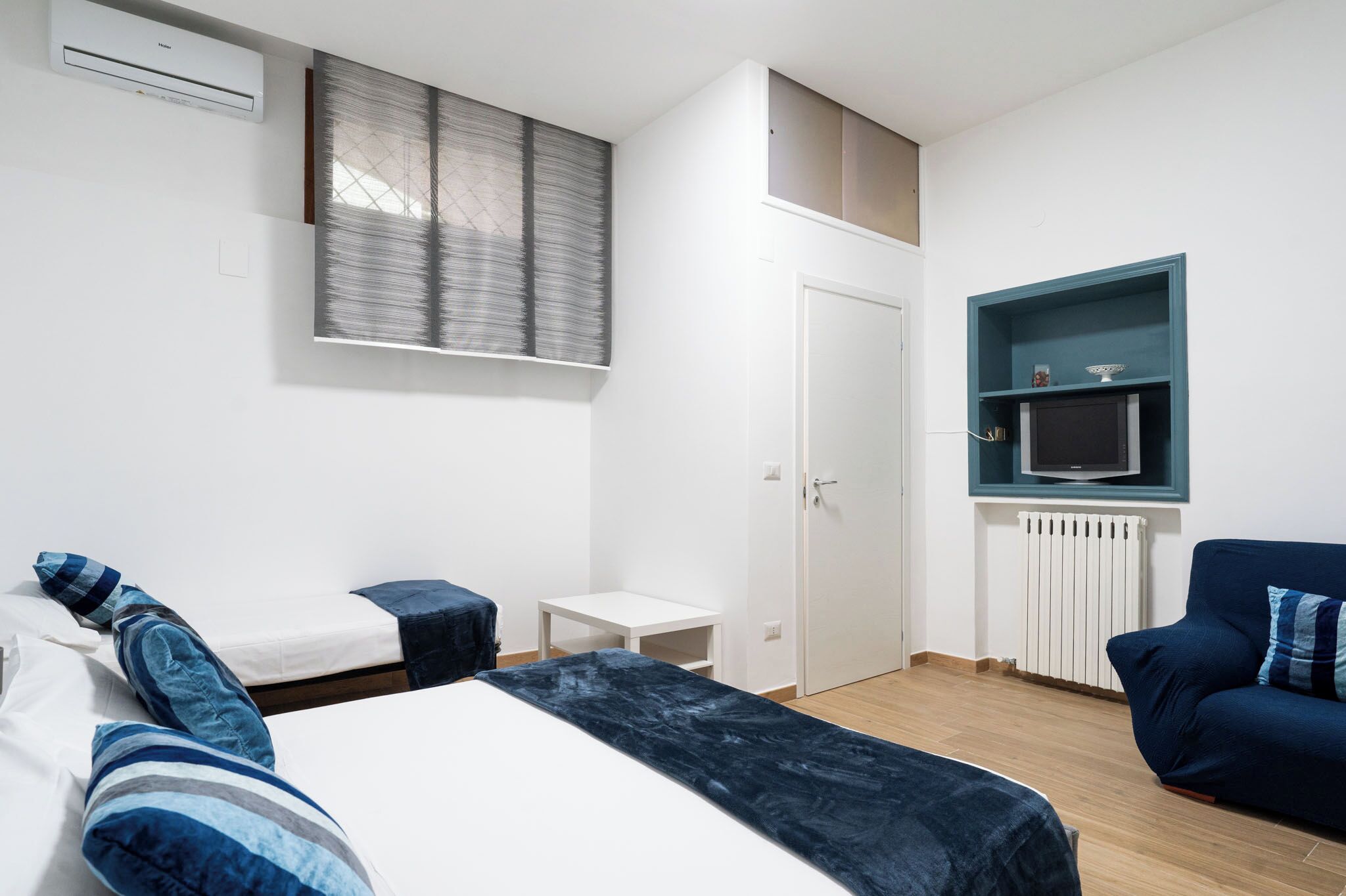 Quarto de hotel moderno com cama, uma mesinha, uma TV em uma prateleira e uma porta.
