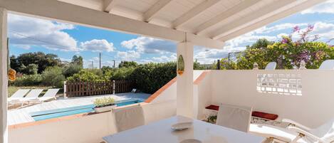 A covered patio area with a table and chairs, a view of a swimming pool, and a wooden fence.