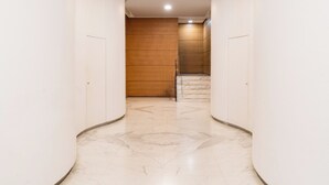 Un pasillo con paredes blancas y piso de mármol, que lleva a una puerta de madera y una escalera.