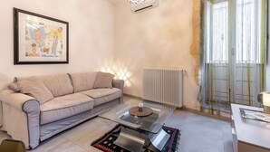 A living room with a sofa, a glass coffee table, a framed picture on the wall, a radiator, and a window with curtains. - Nicu Amuri con Terrazza a Noto by Wonderful Italy (Noto)
