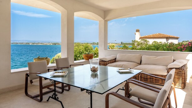 A sunlit room with a glass table, chairs, and a sofa facing a scenic view of the sea and a building. - Villa Plemmiria by the Sea by Wonderful Italy (Syracuse)