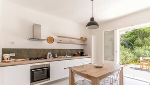 A modern kitchen with a wooden table and chairs, a built-in oven, a gas stove, and a large window offering a view of greenery. - Villa Plemmiria by the Sea by Wonderful Italy (Syracuse)