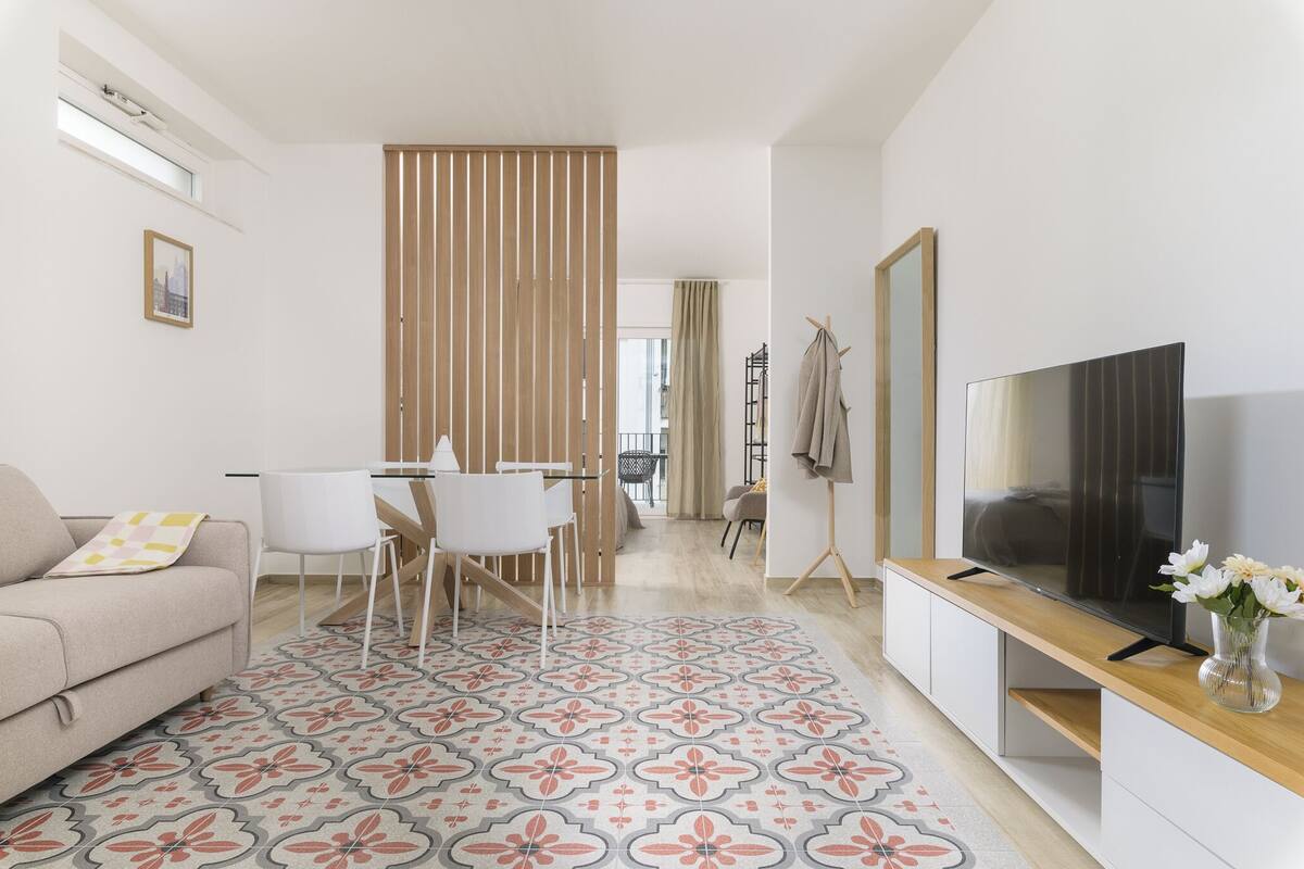 Een moderne woonkamer met een bank, een eettafel met stoelen, een televisie op een dressoir en een kamerkleed met een patroon.