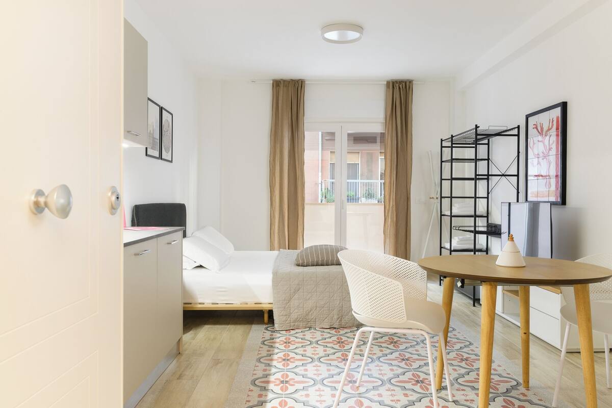 Een kleine hotelkamer met een bed, een bureau, een stoel, een tafeltje en een raam met gordijnen.