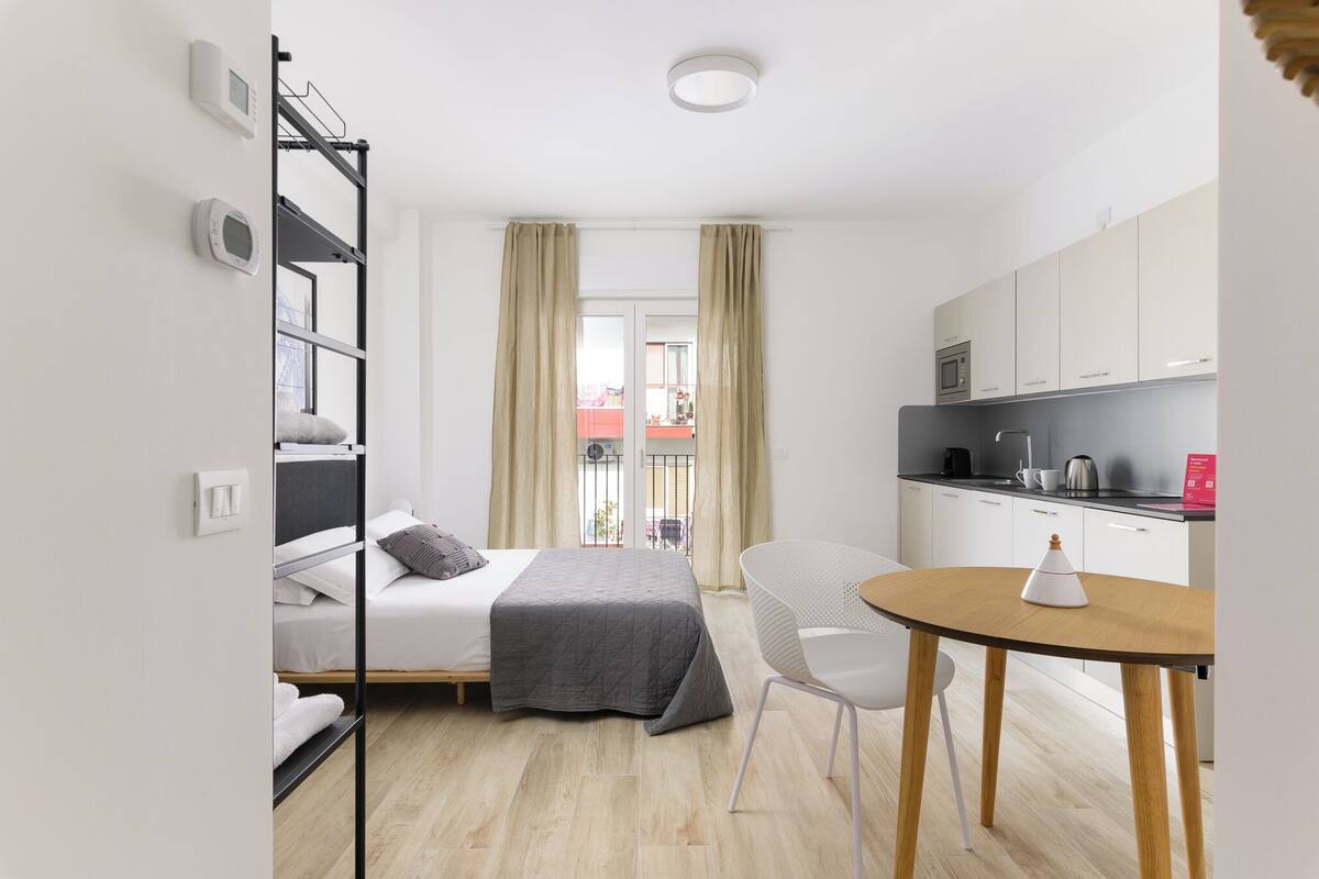 Een compacte hotelkamer met een bed, een tafeltje, een stoel, een kitchenette en een balkon met gordijnen.