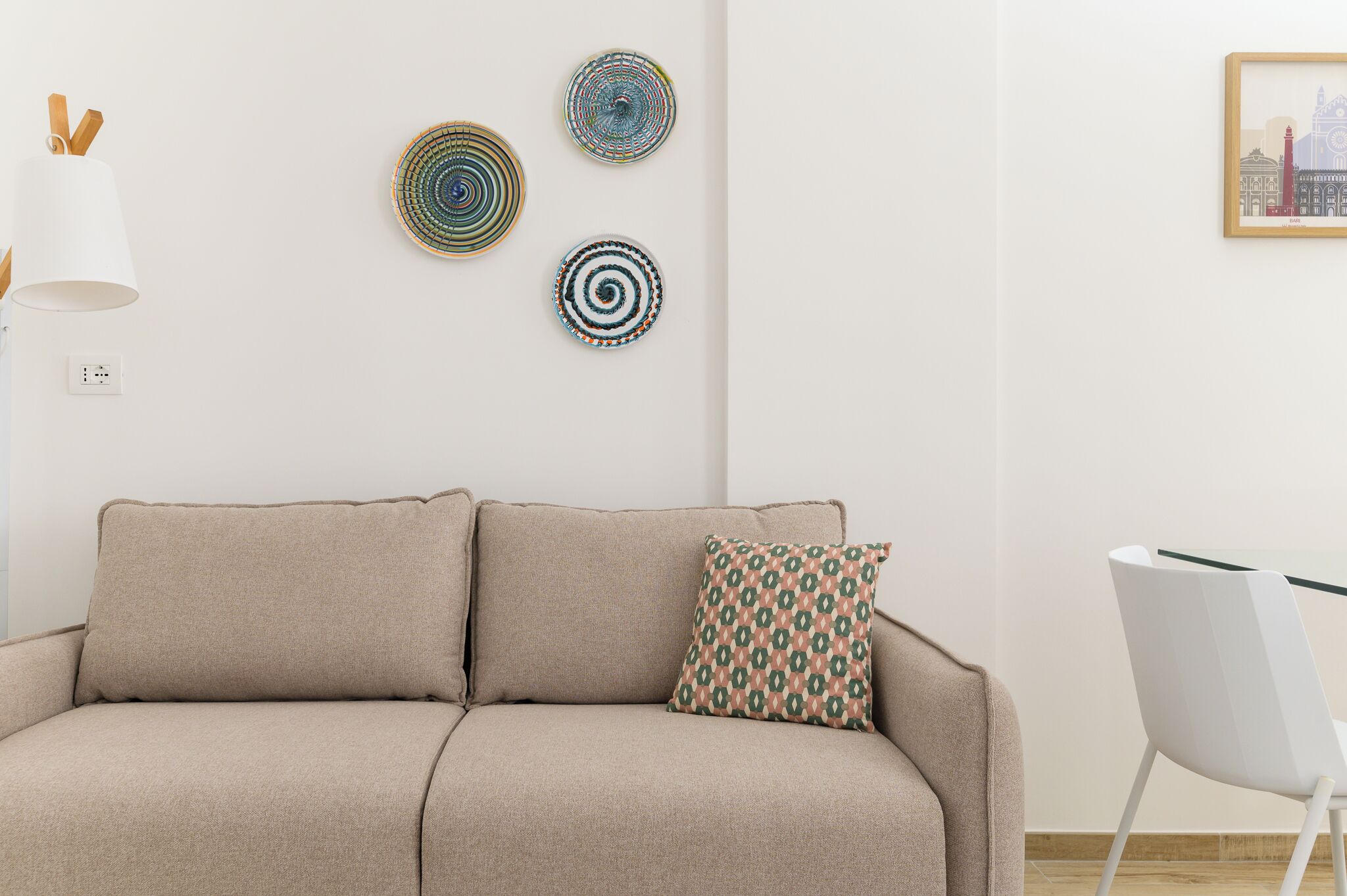 A beige sofa with a patterned pillow, a white lamp, and three wall art pieces.