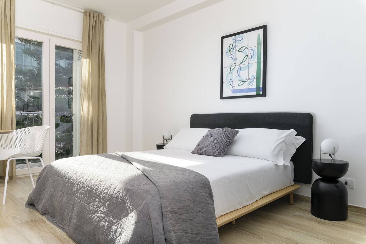 Een moderne slaapkamer met een bed, nachtkastjes, een stoel en een groot raam met gordijnen.
