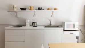 Una cocina moderna con armarios blancos, una encimera blanca, una cafetera, un hervidor, un microondas y una repisa con tazas y cuencos variados.