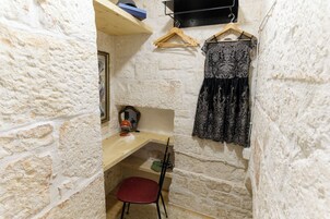 Een kleine kamer met stenen muren, een houten bureau, een rode stoel, en een zwarte kantjurk hangt aan de muur.