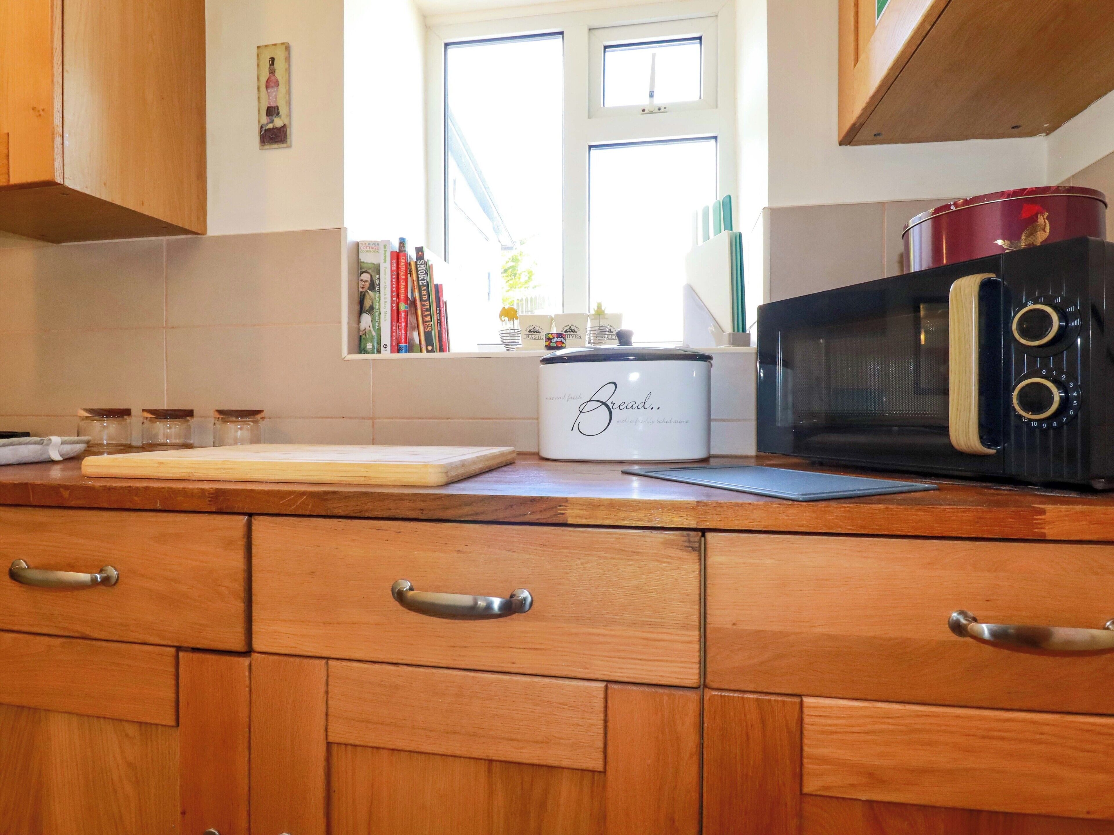 A kitchen with wooden cabinets, a bread maker, a microwave, and a window with books on the windowsill.