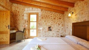 Um quarto rústico com parede de pedra, vigas de madeira no teto, cama com roupa branca, escrivaninha de madeira e uma pequena janela.
